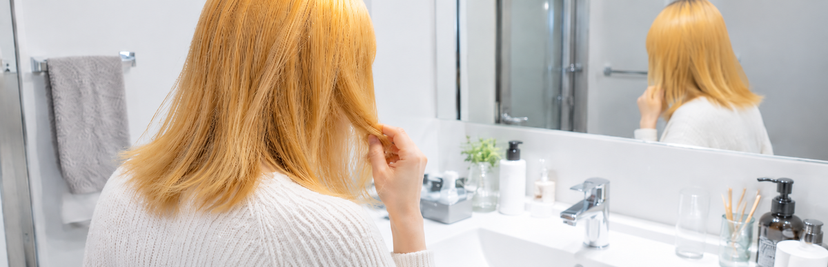 洗面所の白い照明の下で、イエローオレンジっぽい金髪の黄色っぽさを気にする女性の斜め後ろ姿