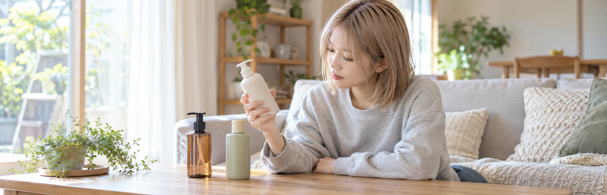リビングのソファでテーブルに並べたトリートメントのボトルを見比べるシルバーベージュのボブの女性