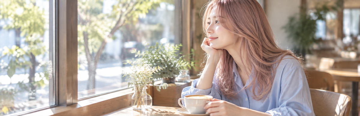 カフェの窓際の席でラテを前にほほえむ、つやのあるピンクベージュの髪の女性