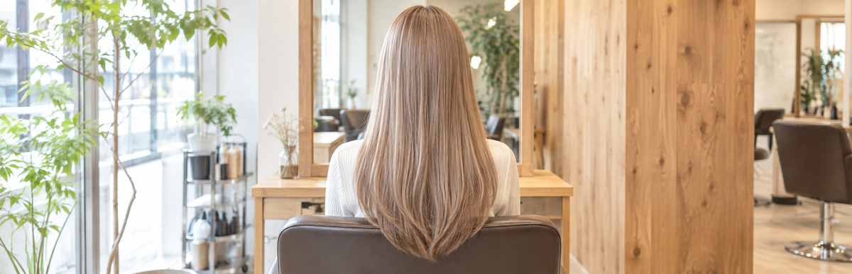 美容室の鏡の前に座る、地毛とエクステの色がそろったミルクティーベージュのロングヘアの後ろ姿