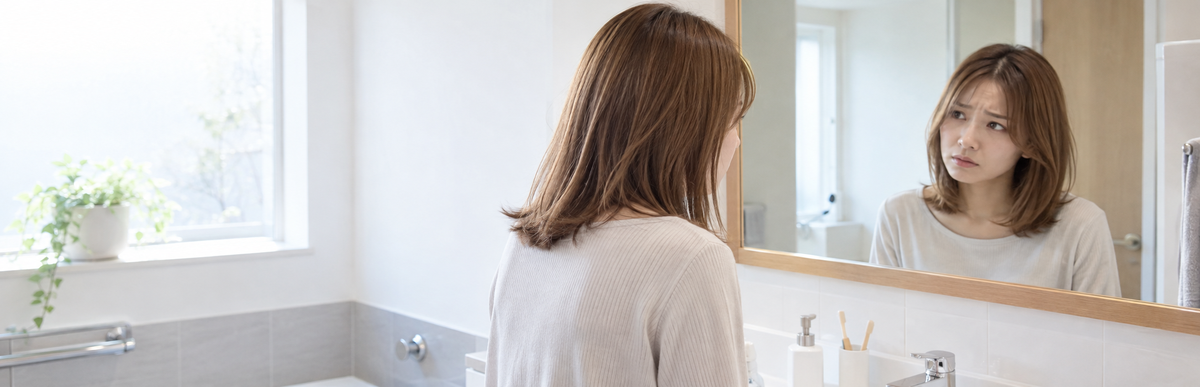 自宅の洗面所の鏡に映った明るめの茶髪を困った表情で見ている女性