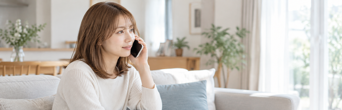 明るいリビングのソファでスマートフォンを耳に当てて美容室に電話している明るい茶髪の女性