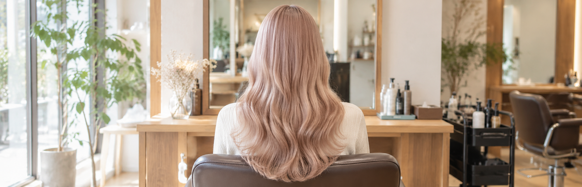 明るい美容室の鏡の前に座る、染めたての白っぽく淡いミルクティーピンクのウェーブヘアの後ろ姿