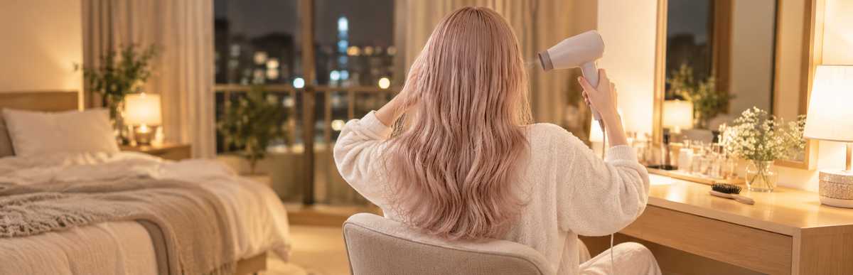 夜の部屋で、シャンプー後の淡いミルクティーピンクの髪をドライヤーで乾かしている女性の後ろ姿