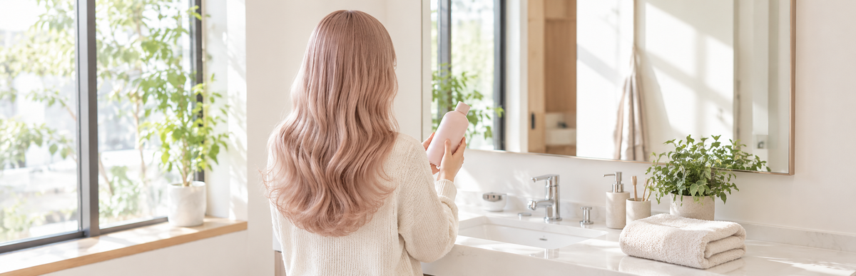 明るい洗面所で淡いピンクのシャンプーボトルを手に持つ、ミルクティーピンクのウェーブヘアの女性の後ろ姿