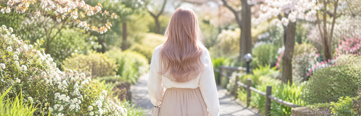 花の咲く春の公園の小道を歩く、白っぽく淡いミルクティーピンクのロングヘアの女性の後ろ姿